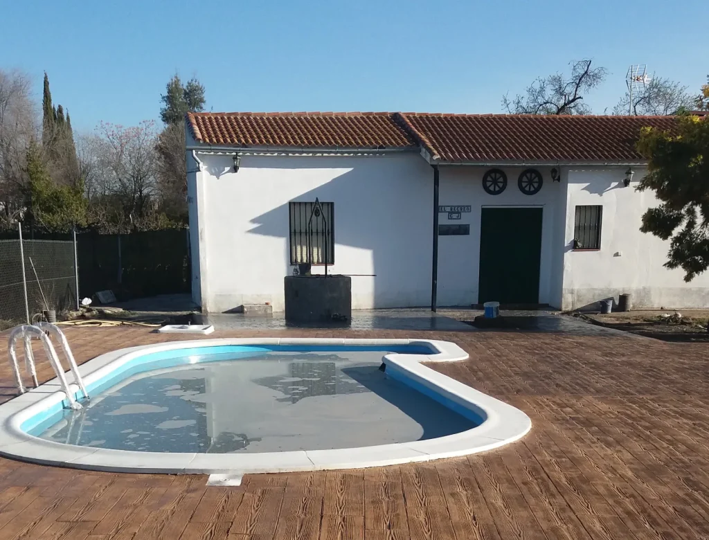 Una terraza de madera con vistas a una piscina en el patio trasero, rodeada de vegetación y luz solar.